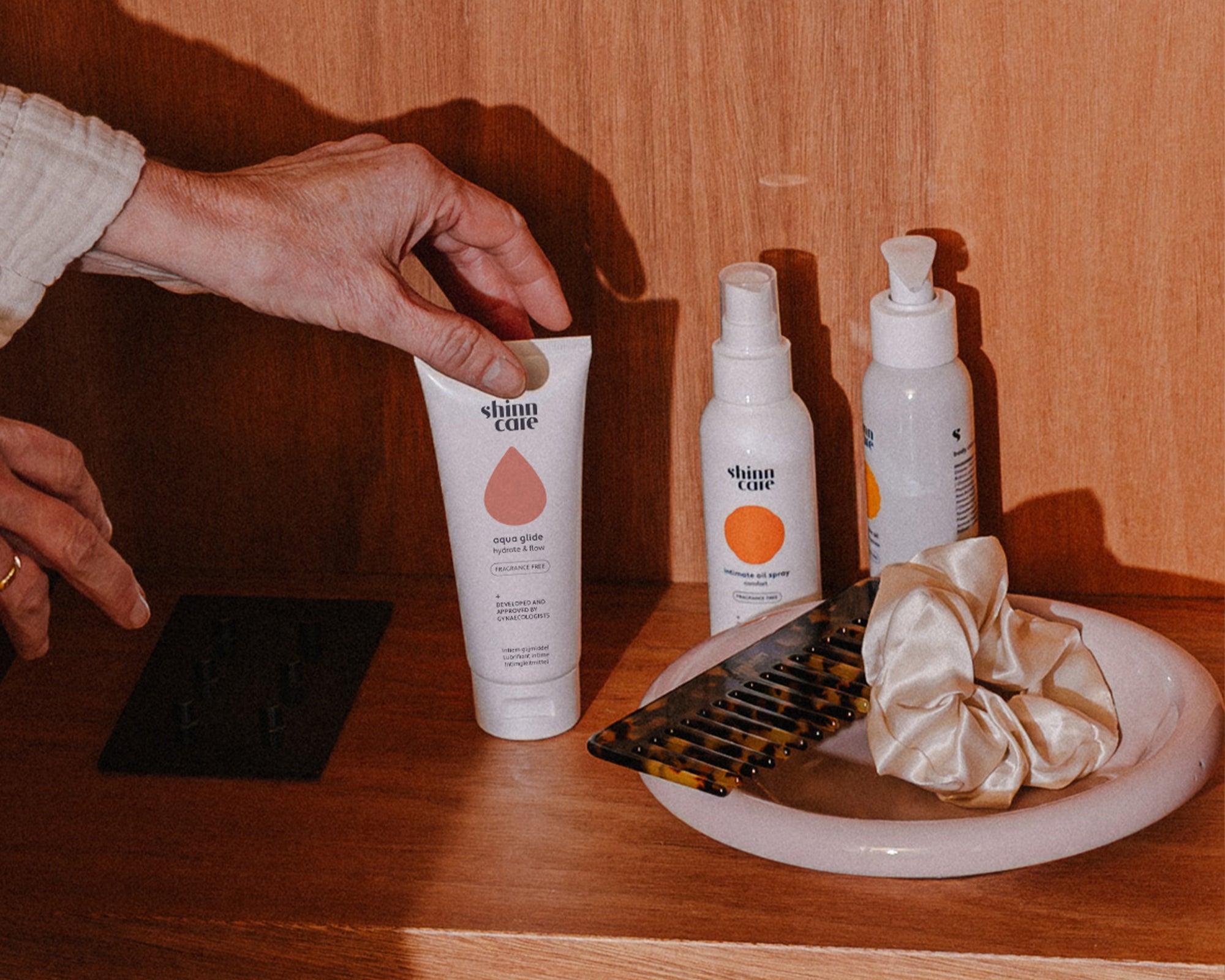Une main se tend vers « Aqua Glide » à côté de « Intimate Oil Spray » Confort sur une table de chevet