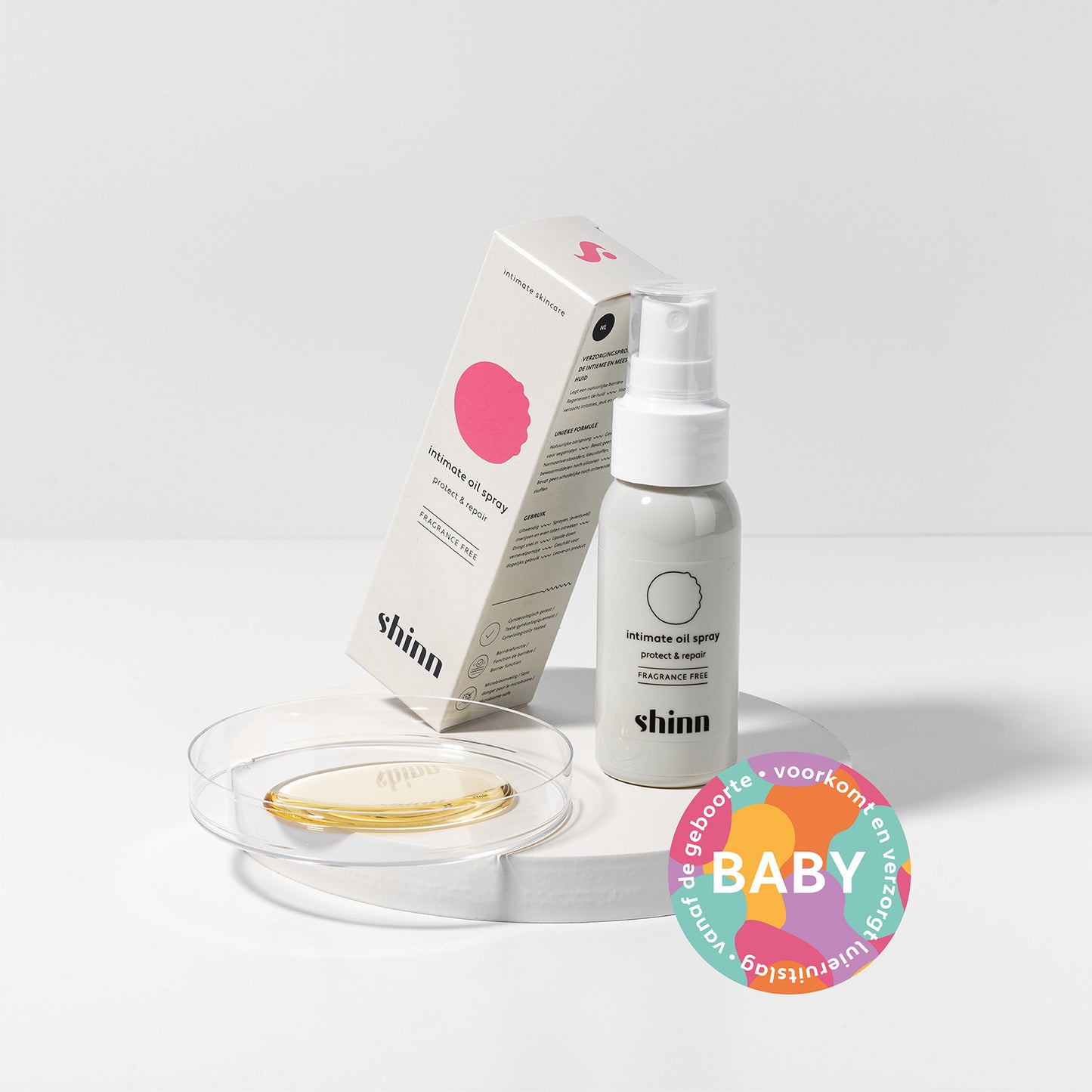 Afbeelding van product (Intimate Oil Spray - Protect & Repair) en de verpakkingsdoos op een witte achtergrond met een label dat dit product ook geschikt is voor baby's. Het product wordt gecombineerd met een petrischaaltje waar het product op werd aangebracht. De Intimate Oil Spray - Protect & Repair - is een droge olie die een natuurlijke bescherming legt op de huid tegen vocht, wrijving of irriterend materiaal.